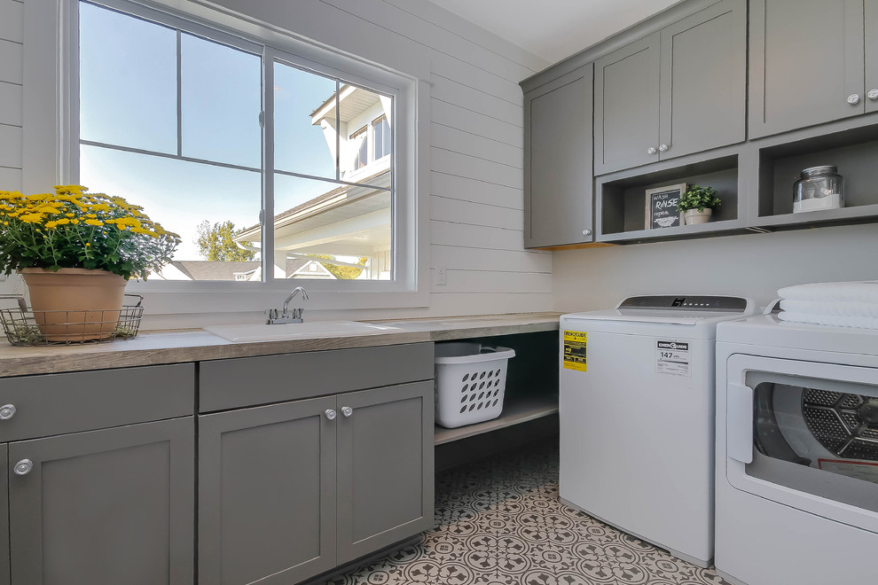Mid-sized country l-shaped dedicated laundry room in Grand Rapids with a drop-in sink, shaker cabinets, grey cabinets, laminate benchtops, white walls, vinyl floors, a side-by-side washer and dryer, grey floor and brown benchtop.
