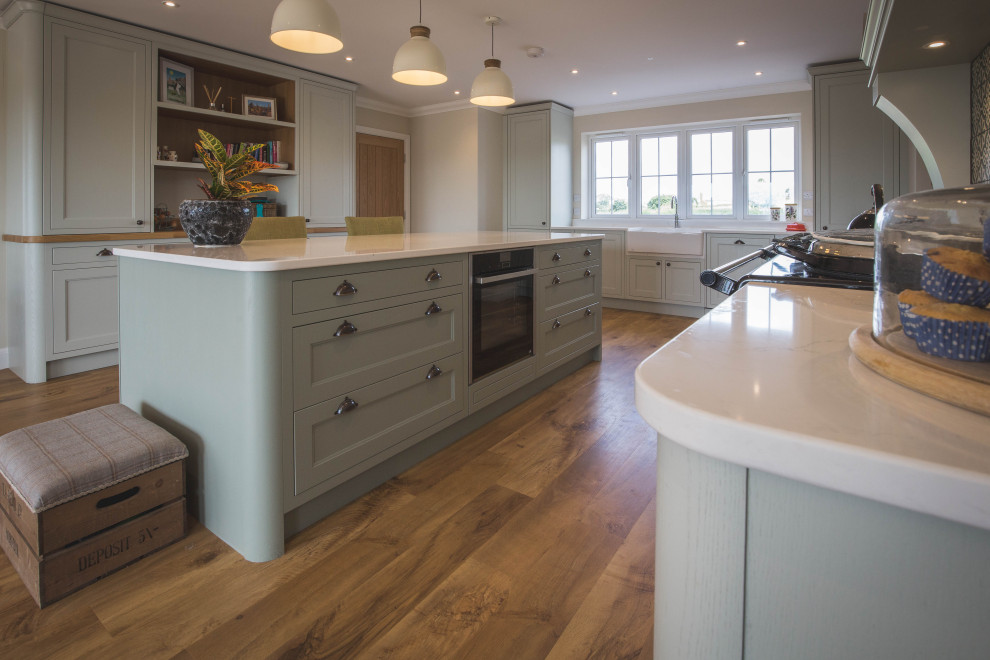 Design ideas for a large classic u-shaped open plan kitchen in Devon with a belfast sink, beaded cabinets, green cabinets, quartz worktops, black appliances, medium hardwood flooring, an island, brown floors and white worktops.