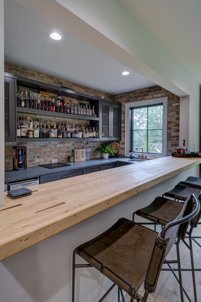 Photo of a mid-sized industrial l-shaped seated home bar in Atlanta with an undermount sink, shaker cabinets, black cabinets, granite benchtops, multi-coloured splashback, brick splashback, light hardwood floors, brown floor and black benchtop.