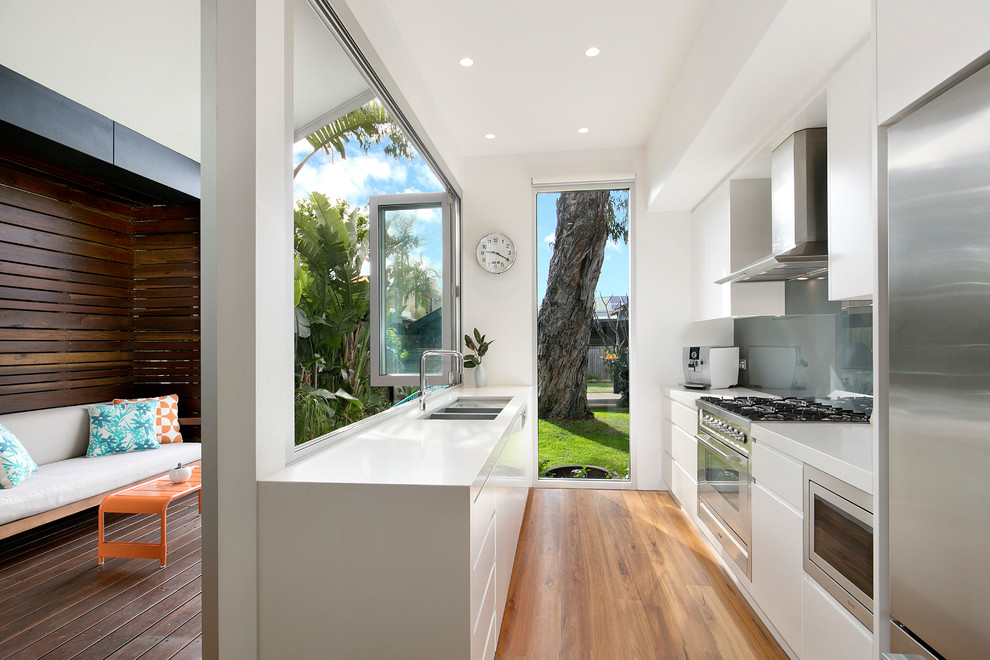 Design ideas for a contemporary galley eat-in kitchen in Sydney with a double-bowl sink, white cabinets, glass sheet splashback, stainless steel appliances, flat-panel cabinets, medium hardwood floors, no island, brown floor, grey splashback and white benchtop.