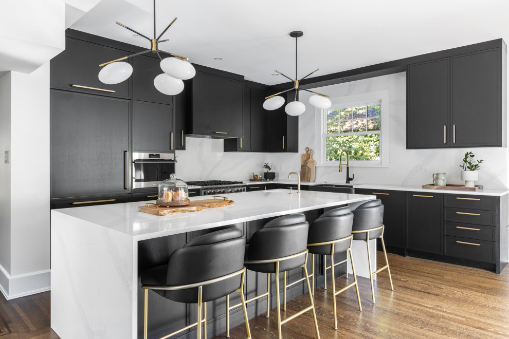 Photo of a contemporary l-shaped kitchen/diner in Vancouver with shaker cabinets, black cabinets, engineered stone countertops, white splashback, engineered quartz splashback, stainless steel appliances, an island, brown floors, white worktops, a belfast sink and medium hardwood flooring.