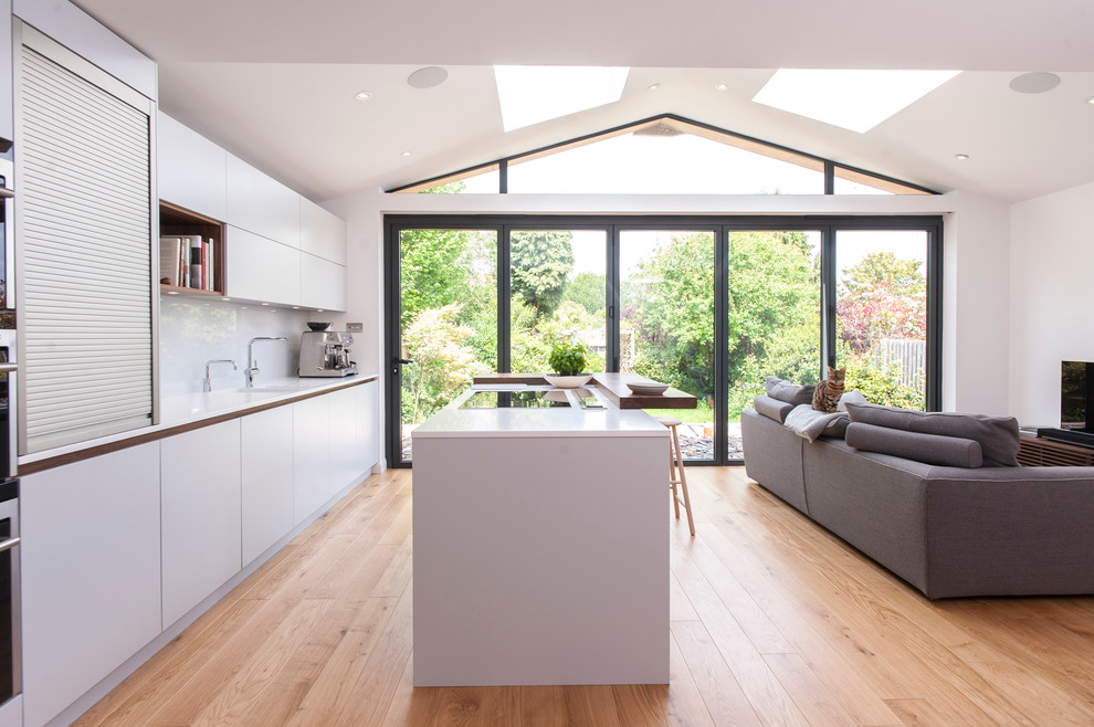 This is an example of a contemporary galley open plan kitchen in London with flat-panel cabinets, white cabinets, white splashback, light hardwood floors and with island.