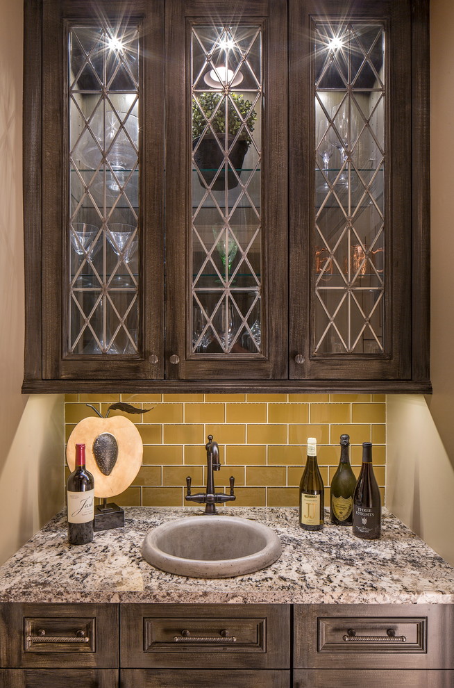 Small transitional single-wall wet bar in Omaha with a drop-in sink, glass-front cabinets, dark wood cabinets, granite benchtops, beige splashback and glass tile splashback.