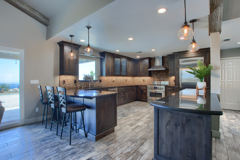 Example of a large mountain style l-shaped porcelain tile, brown floor and vaulted ceiling open concept kitchen design with an undermount sink, shaker cabinets, brown cabinets, granite countertops, brown backsplash, marble backsplash, stainless steel appliances, an island and black countertops