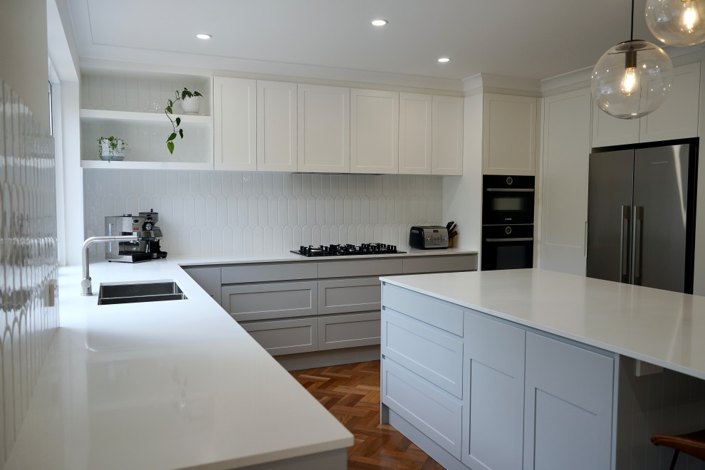 Inspiration for a mid-sized transitional eat-in kitchen in Sydney with an undermount sink, shaker cabinets, grey cabinets, quartz benchtops, white splashback, ceramic splashback, black appliances, medium hardwood floors, with island, brown floor and white benchtop.