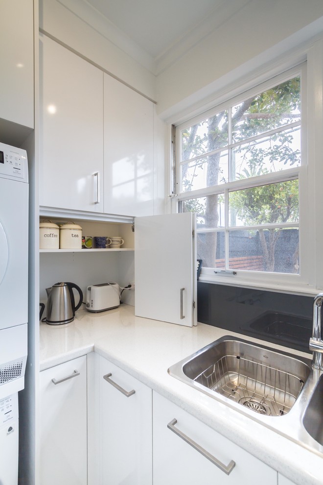 This is an example of a small modern l-shaped utility room in Melbourne with a double-bowl sink, flat-panel cabinets, white cabinets, laminate benchtops, white walls, light hardwood floors and a stacked washer and dryer.