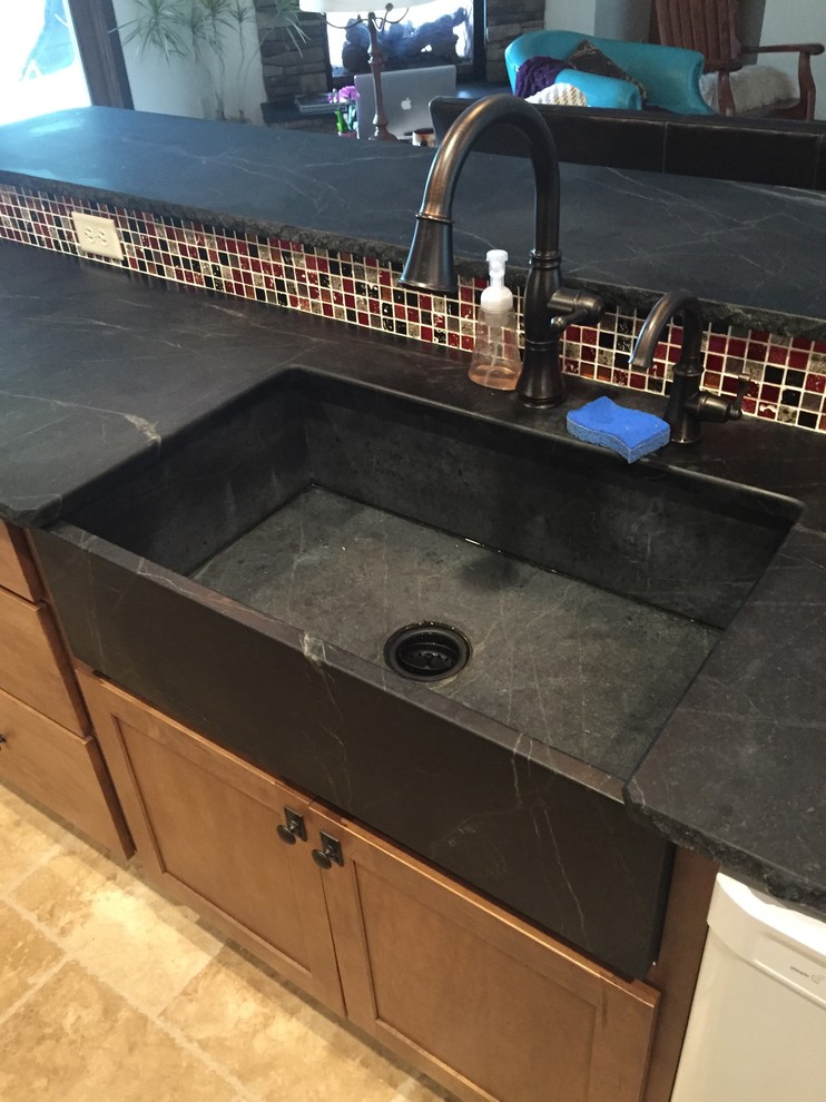 Large mountain style galley travertine floor open concept kitchen photo in Columbus with a farmhouse sink, shaker cabinets, medium tone wood cabinets, soapstone countertops, beige backsplash, stone tile backsplash, white appliances and an island
