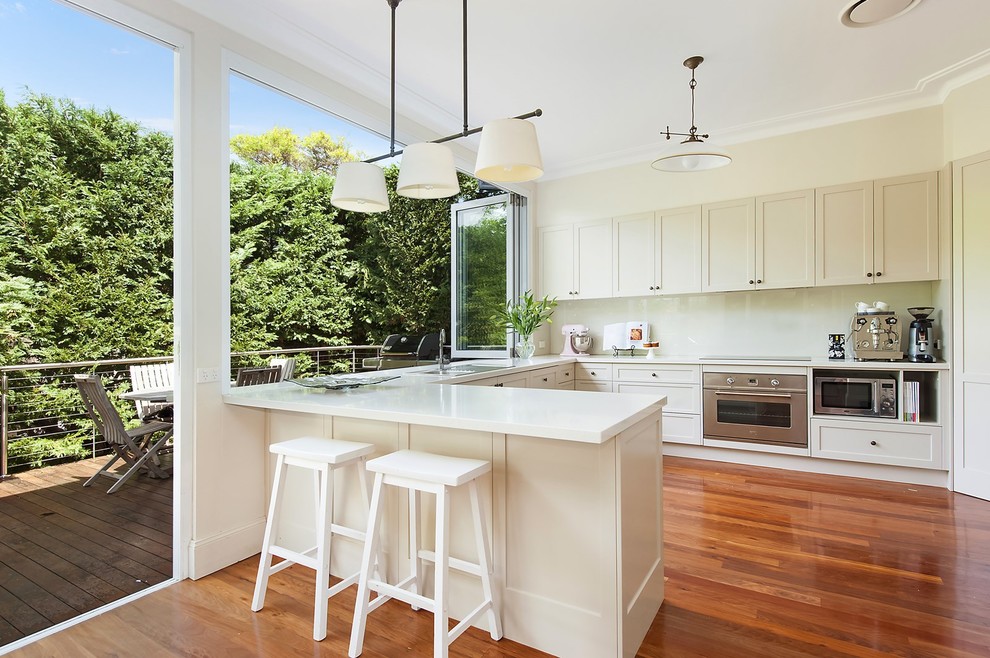 Design ideas for a transitional u-shaped kitchen in Sydney with a drop-in sink, shaker cabinets, white cabinets, white splashback, glass sheet splashback, stainless steel appliances, medium hardwood floors, a peninsula, brown floor and white benchtop.
