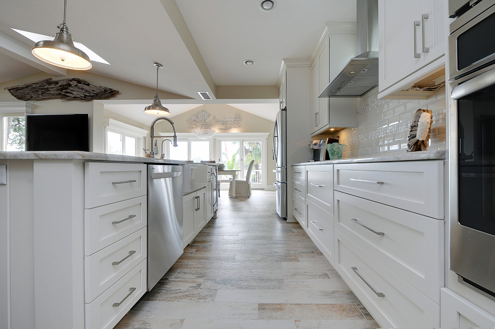 Design ideas for a mid-sized beach style single-wall open plan kitchen in Tampa with a farmhouse sink, shaker cabinets, white cabinets, granite benchtops, white splashback, ceramic splashback, stainless steel appliances, vinyl floors and with island.