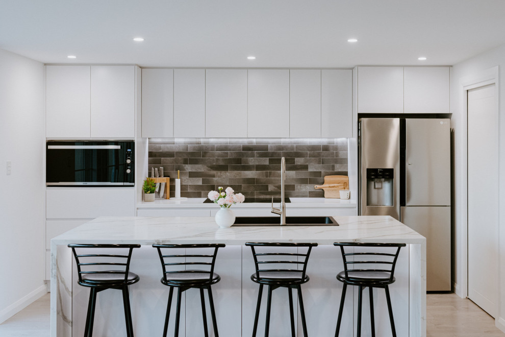 Mid-sized contemporary galley kitchen in Sydney with a drop-in sink, flat-panel cabinets, white cabinets, grey splashback, stainless steel appliances, with island, beige floor and grey benchtop.