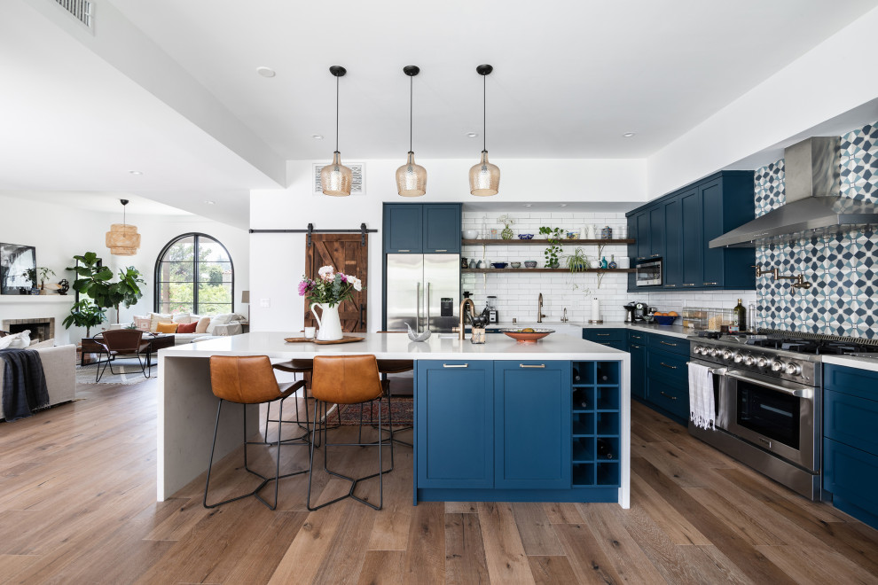 This is an example of a mid-sized mediterranean l-shaped eat-in kitchen in Los Angeles with a farmhouse sink, shaker cabinets, blue cabinets, quartz benchtops, white splashback, ceramic splashback, stainless steel appliances, medium hardwood floors, with island, brown floor and white benchtop.