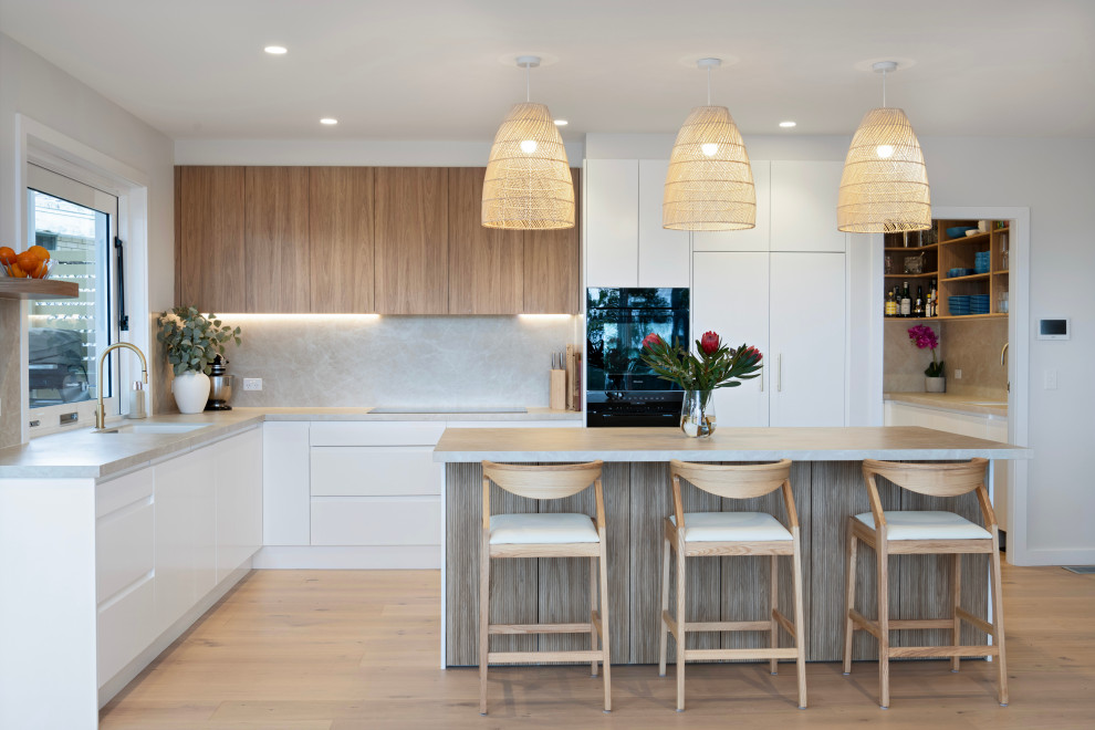 This is an example of a modern l-shaped kitchen in Sydney with an undermount sink, flat-panel cabinets, white cabinets, beige splashback, stone slab splashback, black appliances, light hardwood floors, with island, beige floor and beige benchtop.