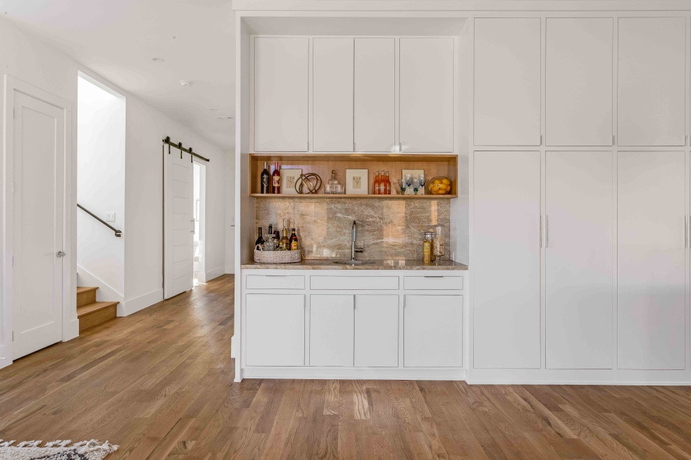 Photo of a large beach style single-wall wet bar in Dallas with an undermount sink, flat-panel cabinets, white cabinets, quartzite benchtops, brown splashback, stone slab splashback, light hardwood floors, brown floor and brown benchtop.