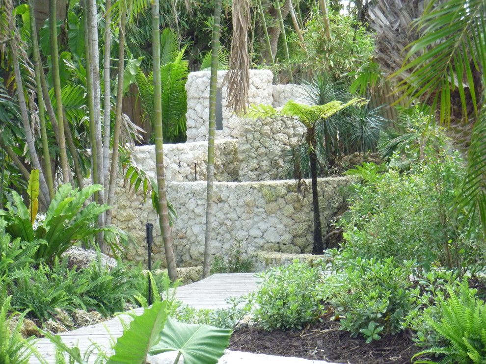 Foto di un grande giardino tropicale in ombra dietro casa con un muro di contenimento e pavimentazioni in pietra naturale
