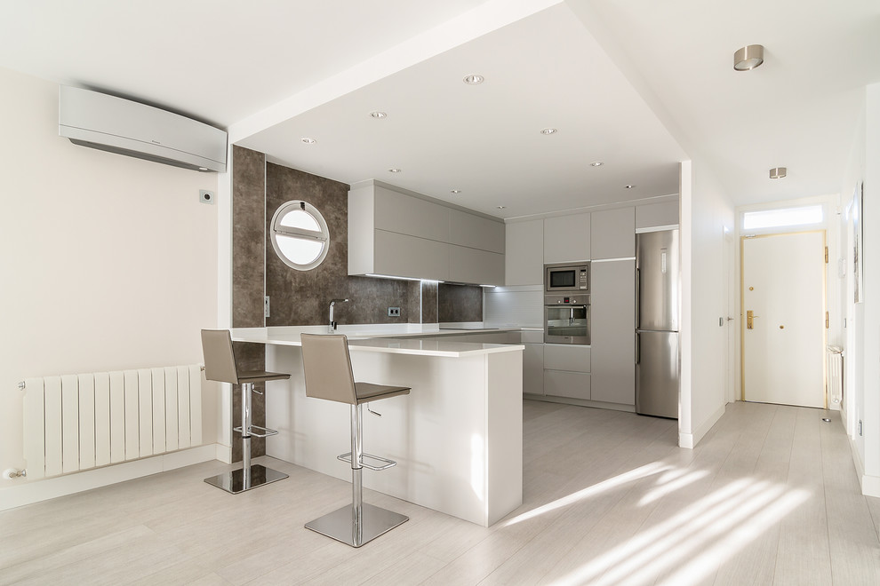 This is an example of a contemporary u-shaped kitchen in Madrid with grey cabinets, light hardwood floors, a peninsula, white benchtop, flat-panel cabinets, brown splashback, stainless steel appliances and grey floor.