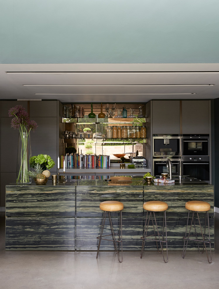 This is an example of a contemporary galley kitchen in London with flat-panel cabinets, grey cabinets, metallic splashback, mirror splashback, stainless steel appliances, concrete floors, with island and green benchtop.