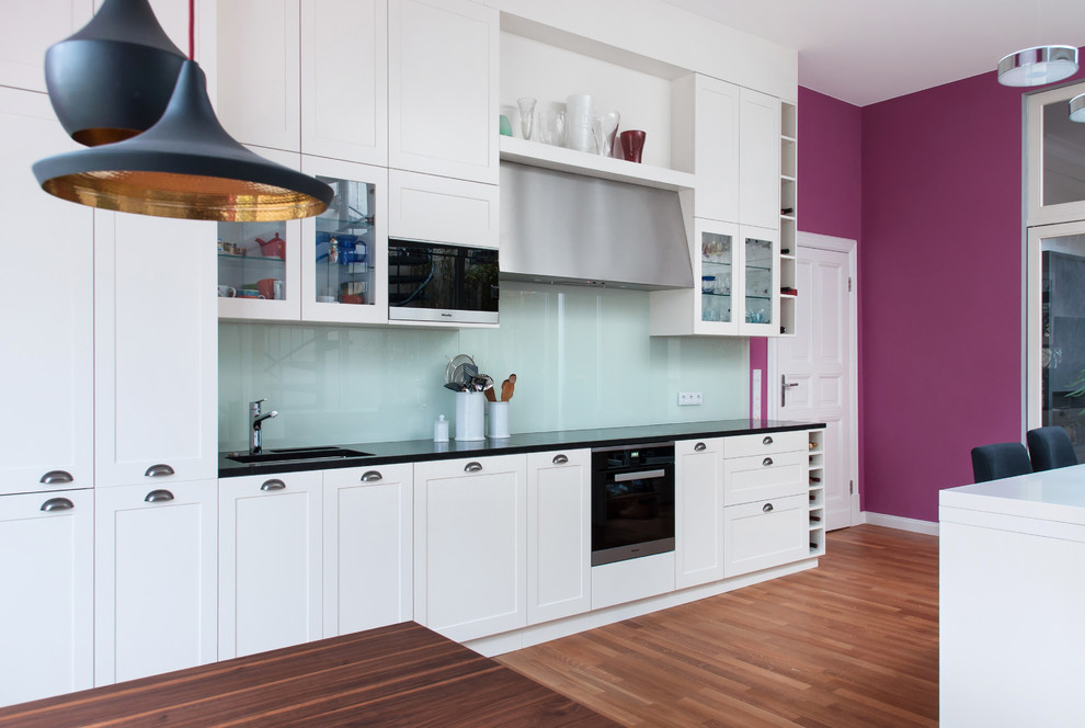 This is an example of a large contemporary kitchen in Berlin with shaker cabinets, white cabinets, blue splashback, glass sheet splashback, black appliances, medium hardwood floors and brown floor.
