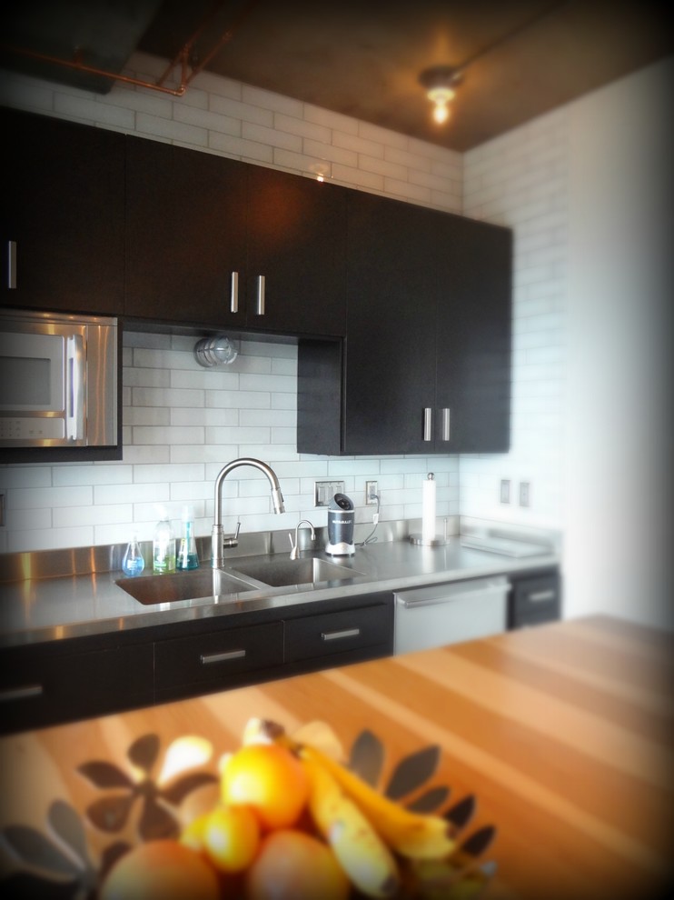 Photo of a mid-sized industrial single-wall eat-in kitchen in Portland with an integrated sink, flat-panel cabinets, stainless steel benchtops, white splashback, stainless steel appliances, concrete floors, with island, black cabinets and subway tile splashback.