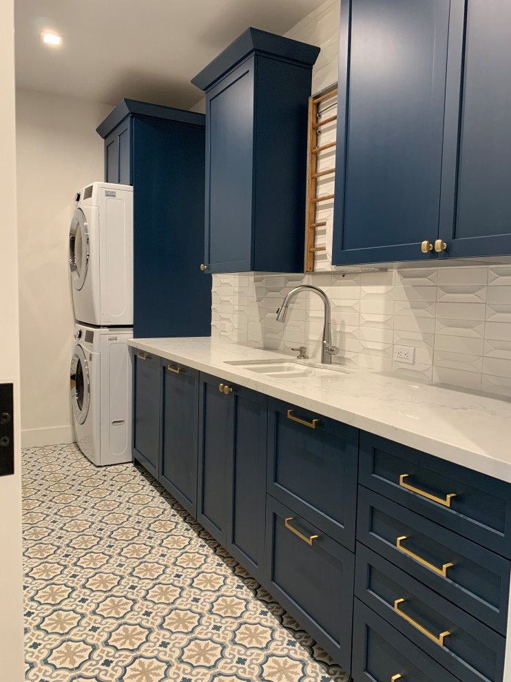 オレンジカウンティにある高級な中くらいなビーチスタイルのおしゃれな洗濯室 (ll型、ダブルシンク、シェーカースタイル扉のキャビネット、青いキャビネット、クオーツストーンカウンター、白いキッチンパネル、セラミックタイルのキッチンパネル、白い壁、セラミックタイルの床、上下配置の洗濯機・乾燥機、マルチカラーの床、白いキッチンカウンター) の写真