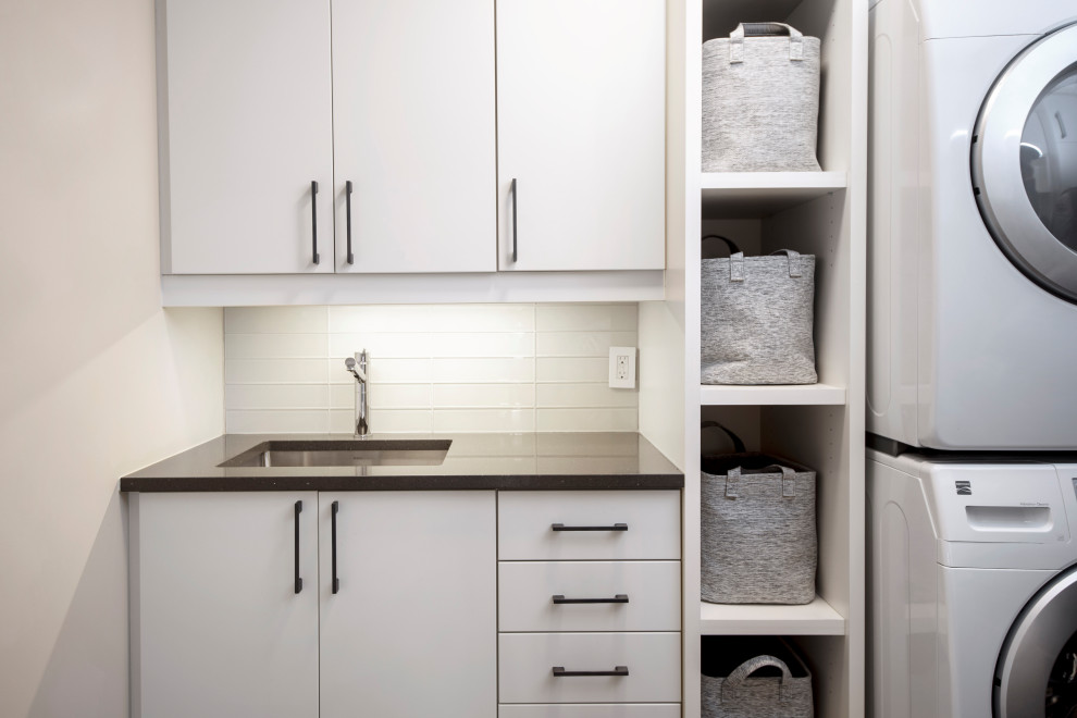 Small modern galley utility room in Ottawa with a single-bowl sink, flat-panel cabinets, white cabinets, quartzite benchtops, white splashback, ceramic splashback, white walls, ceramic floors, a stacked washer and dryer, black benchtop and multi-coloured floor.