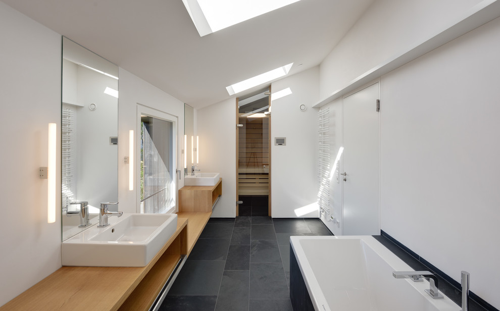 Mid-sized contemporary master bathroom in Berlin with a vessel sink, light wood cabinets, wood benchtops, a drop-in tub, black tile, stone slab, white walls, slate floors and open cabinets.
