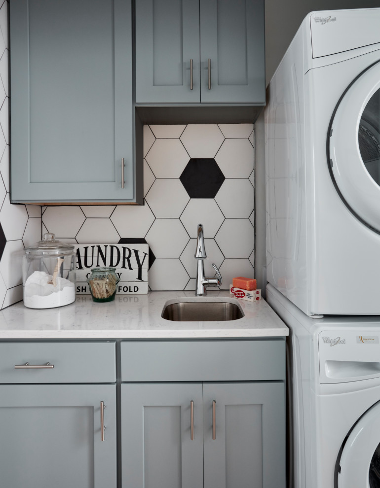 Inspiration for a small transitional single-wall dedicated laundry room in St Louis with an undermount sink, shaker cabinets, blue cabinets, solid surface benchtops, white walls, ceramic floors, a stacked washer and dryer, black floor and white benchtop.