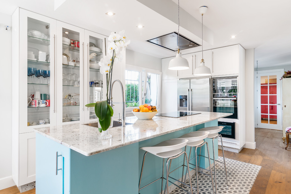 This is an example of a transitional l-shaped kitchen in Other with blue cabinets, stainless steel appliances, with island, a drop-in sink, shaker cabinets and light hardwood floors.