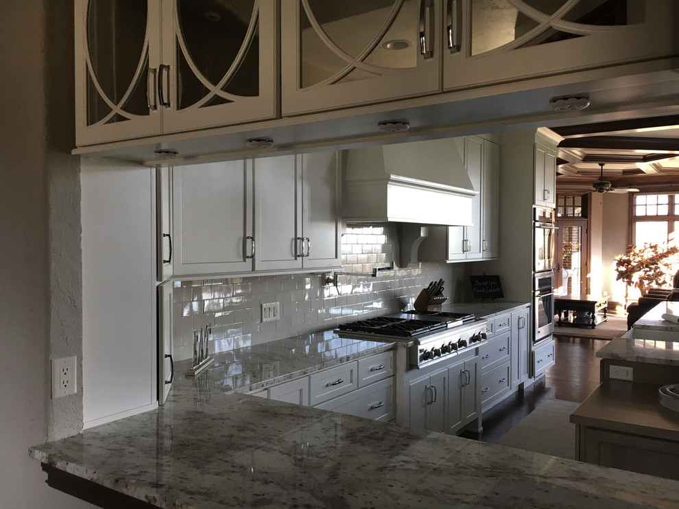 Large transitional l-shaped dark wood floor and brown floor eat-in kitchen photo with a farmhouse sink, recessed-panel cabinets, white cabinets, granite countertops, white backsplash, ceramic backsplash, stainless steel appliances and an island