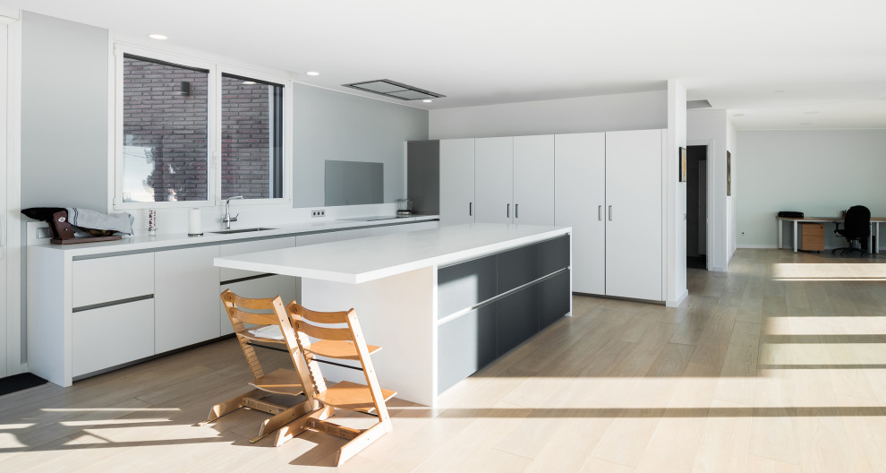 This is an example of a mid-sized modern single-wall open plan kitchen in Barcelona with recessed-panel cabinets, white cabinets, white splashback, light hardwood floors, with island, brown floor and white benchtop.