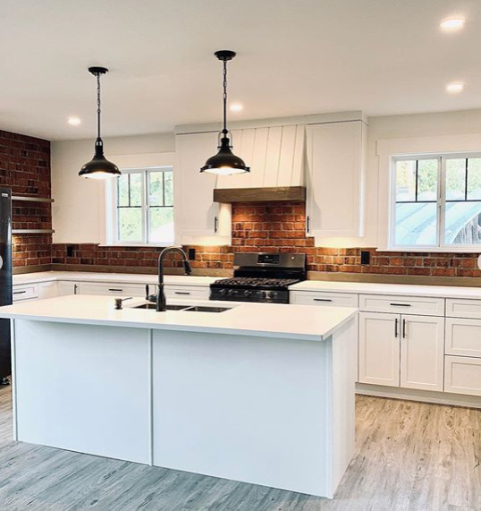 This is an example of a mid-sized country l-shaped kitchen in Vancouver with an undermount sink, shaker cabinets, white cabinets, quartz benchtops, red splashback, brick splashback, black appliances, vinyl floors, with island, beige floor and white benchtop.