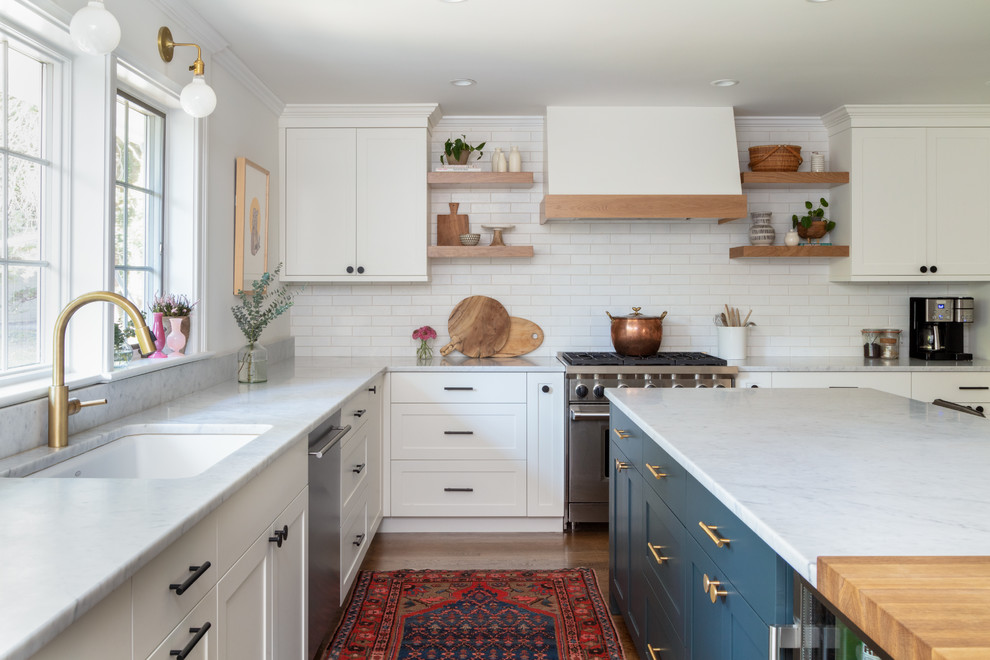 This is an example of a transitional l-shaped kitchen in New York with an undermount sink, shaker cabinets, white cabinets, white splashback, subway tile splashback, stainless steel appliances, medium hardwood floors, with island, brown floor and white benchtop.