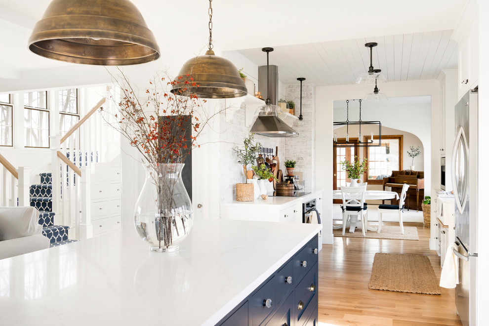 This is an example of a country eat-in kitchen in Minneapolis with a farmhouse sink, white cabinets, brick splashback, stainless steel appliances, light hardwood floors, with island, beige floor and white benchtop.