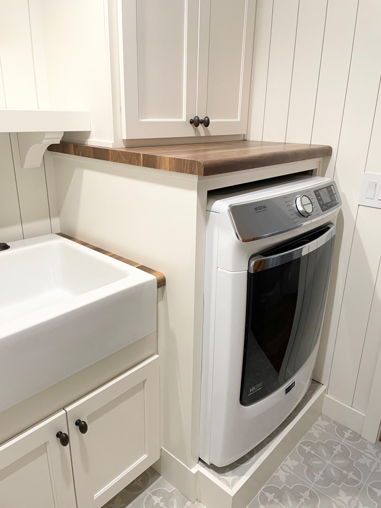 Photo of a mid-sized country single-wall dedicated laundry room in Other with a farmhouse sink, flat-panel cabinets, white cabinets, wood benchtops, beige splashback, shiplap splashback, beige walls, porcelain floors, an integrated washer and dryer, multi-coloured floor, brown benchtop and planked wall panelling.