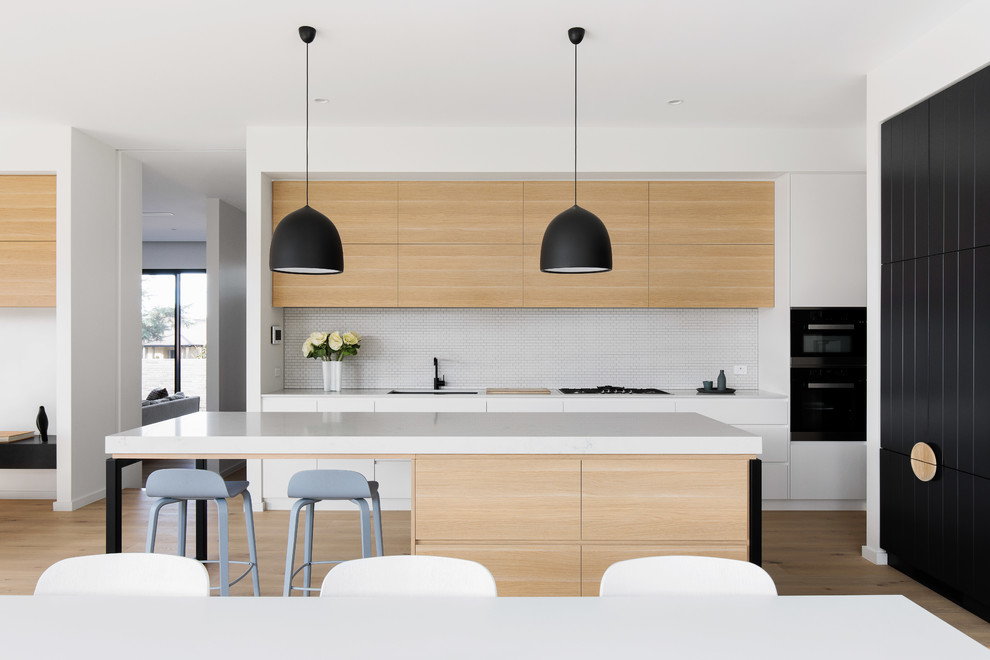Inspiration for a contemporary galley kitchen in Canberra - Queanbeyan with an undermount sink, quartz benchtops, white splashback, mosaic tile splashback, black appliances, light hardwood floors, with island and white benchtop.