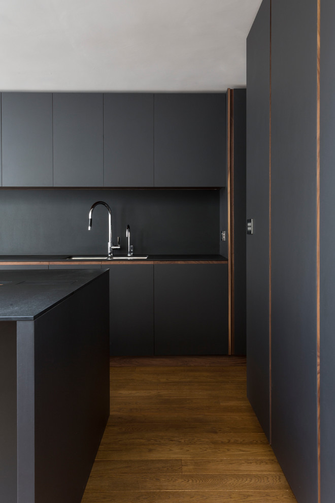 This is an example of a mid-sized contemporary single-wall eat-in kitchen in London with a double-bowl sink, flat-panel cabinets, black cabinets, black splashback, medium hardwood floors, with island and brown floor.
