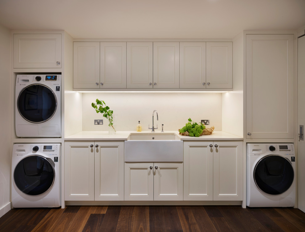 Design ideas for a large traditional single-wall utility room in London with a farmhouse sink, recessed-panel cabinets, white cabinets, solid surface benchtops, white walls, dark hardwood floors, a stacked washer and dryer, brown floor and white benchtop.