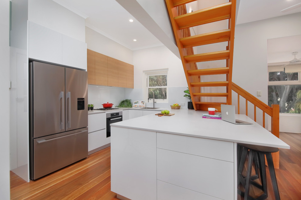Design ideas for a mid-sized contemporary u-shaped open plan kitchen in Sydney with an integrated sink, recessed-panel cabinets, white cabinets, quartz benchtops, white splashback, glass sheet splashback, white appliances, plywood floors, with island, brown floor and white benchtop.