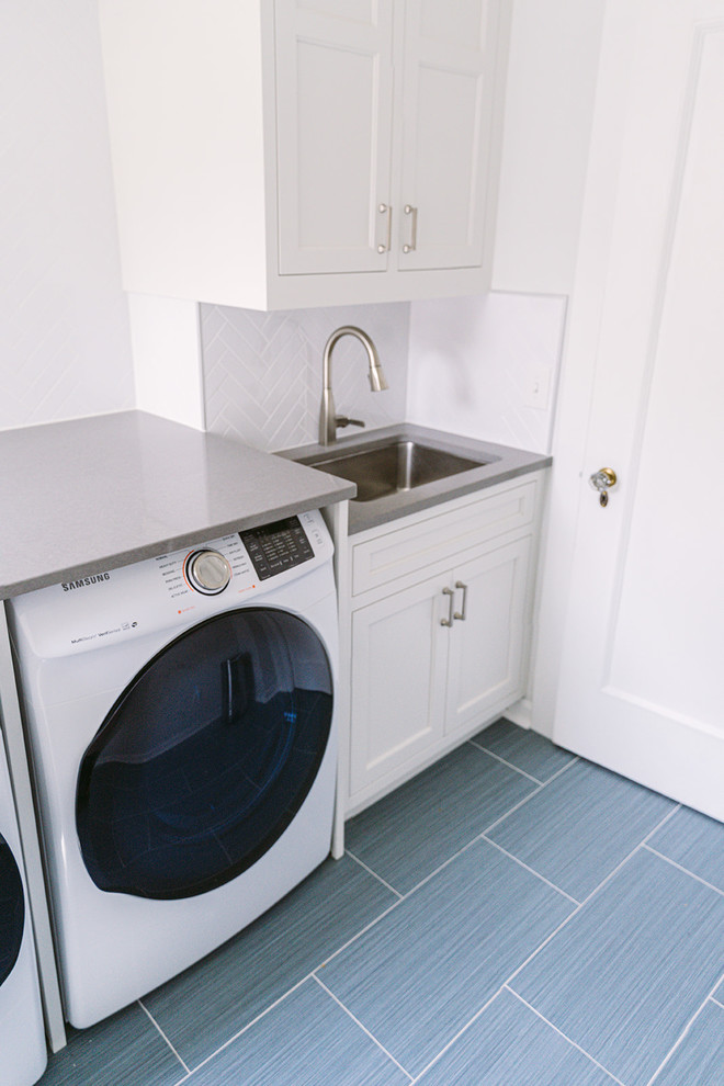 This is an example of a mid-sized transitional galley dedicated laundry room in Cincinnati with a single-bowl sink, white cabinets, quartz benchtops, white walls, ceramic floors, an integrated washer and dryer, grey floor, recessed-panel cabinets and grey benchtop.