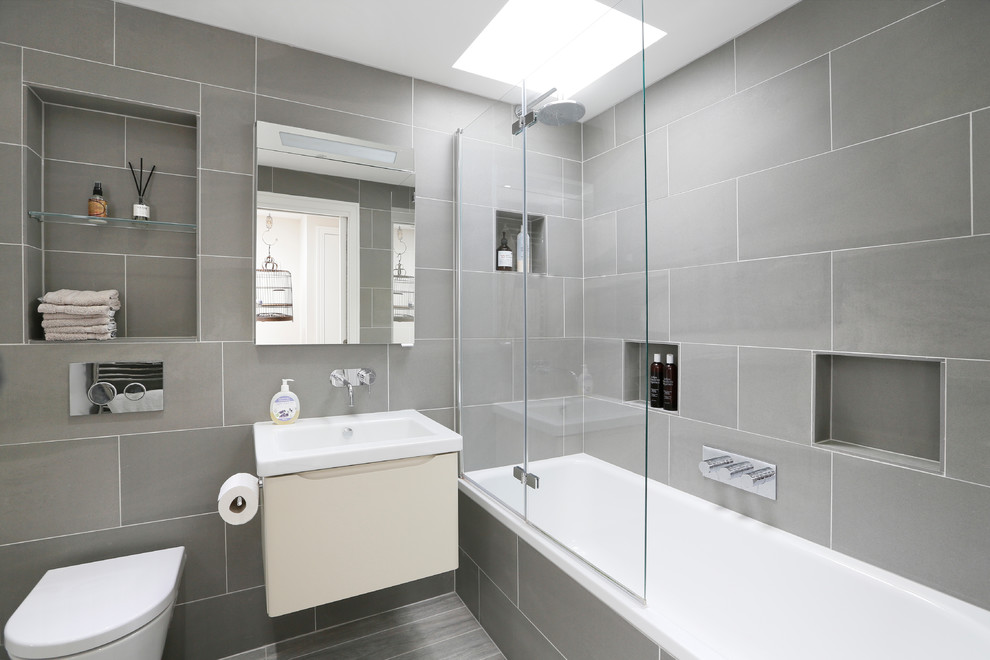 Large contemporary 3/4 bathroom in London with gray tile, flat-panel cabinets, beige cabinets, an alcove tub, a shower/bathtub combo, a wall-mount toilet, grey walls, a wall-mount sink, grey floor, a hinged shower door and white benchtops.