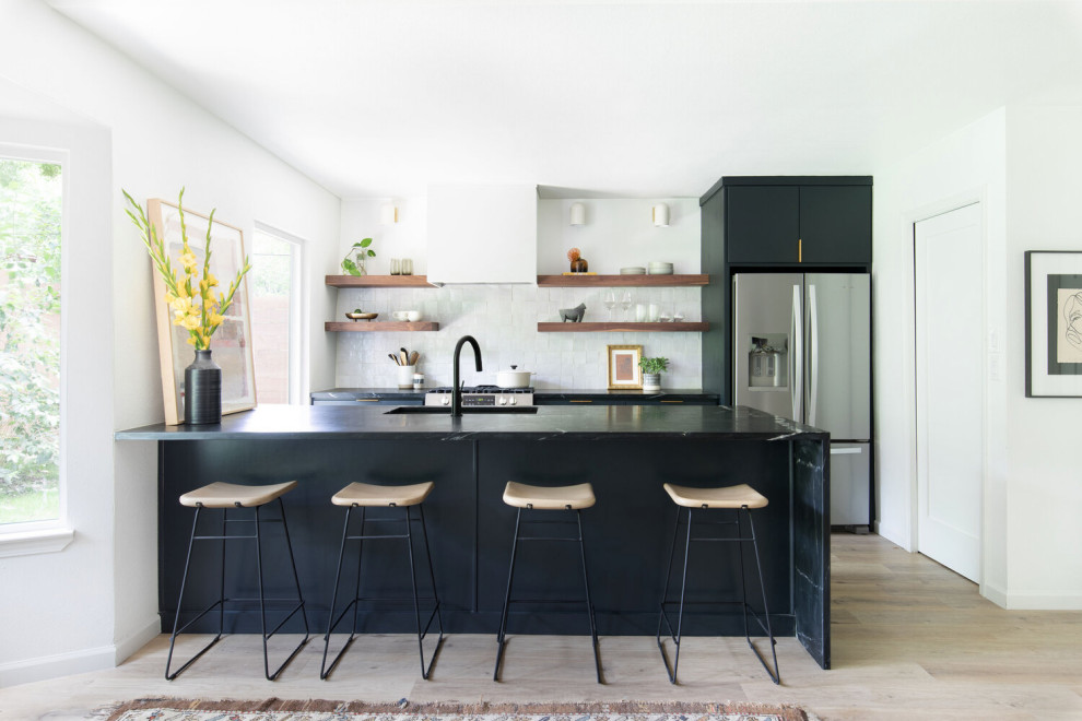 Inspiration for a mid-sized scandinavian galley open plan kitchen in Austin with an undermount sink, flat-panel cabinets, black cabinets, soapstone benchtops, white splashback, ceramic splashback, stainless steel appliances, light hardwood floors, with island, beige floor and black benchtop.