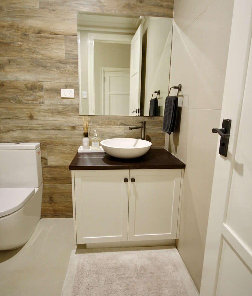 Photo of a mid-sized traditional powder room in Sydney with shaker cabinets, white cabinets, a two-piece toilet, beige tile, ceramic tile, multi-coloured walls, ceramic floors, a vessel sink, wood benchtops, beige floor and brown benchtops.