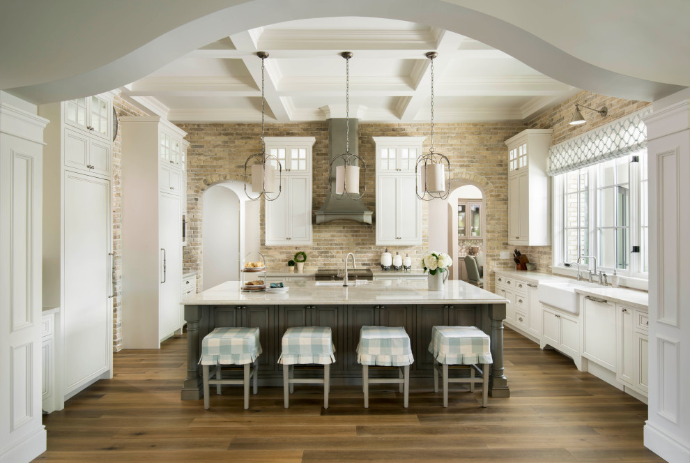 This is an example of an expansive mediterranean u-shaped open plan kitchen in Phoenix with white cabinets, quartzite benchtops, brick splashback, medium hardwood floors, with island, recessed-panel cabinets, a farmhouse sink, grey splashback, panelled appliances, brown floor, white benchtop and coffered.