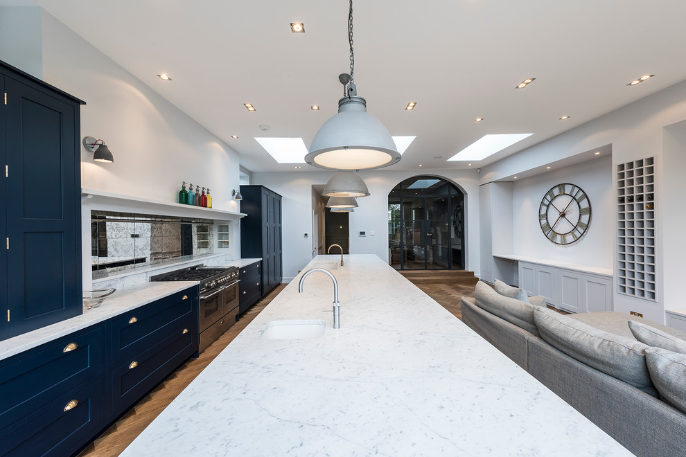 Large transitional open plan kitchen in London with a drop-in sink, shaker cabinets, blue cabinets, marble benchtops, light hardwood floors and with island.