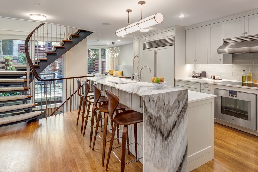 Photo of a transitional kitchen in New York with shaker cabinets, grey cabinets, grey splashback, glass tile splashback, medium hardwood floors, with island and panelled appliances.