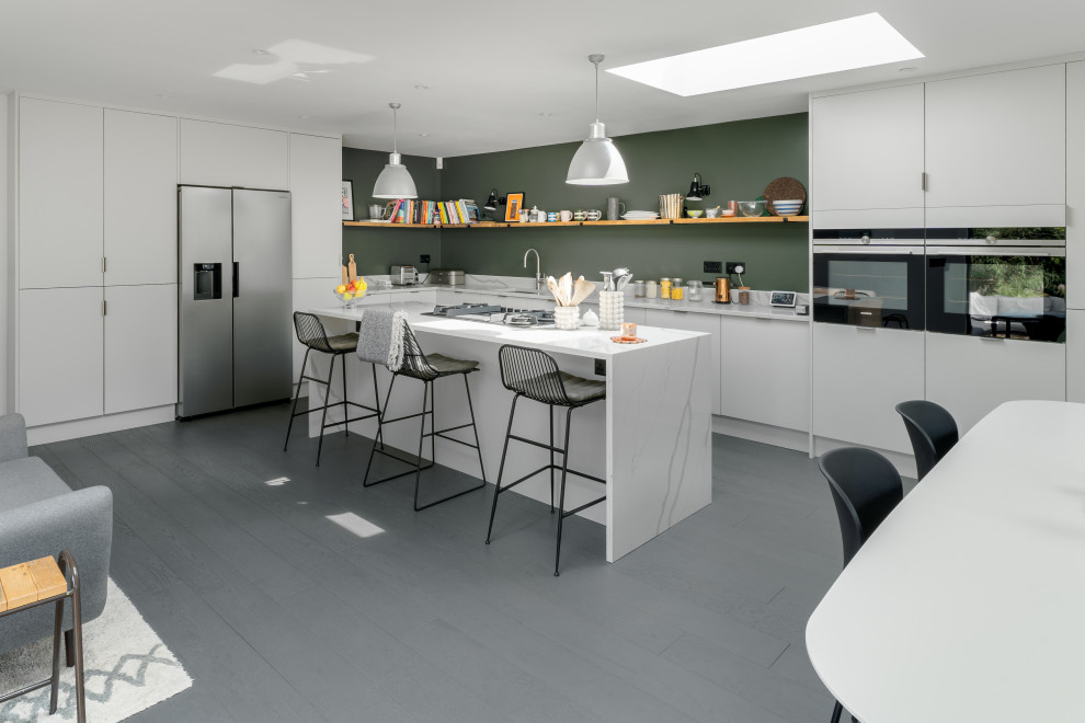 Photo of a medium sized contemporary l-shaped kitchen/diner in Sussex with a submerged sink, flat-panel cabinets, white cabinets, quartz worktops, green splashback, stainless steel appliances, dark hardwood flooring, an island, grey floors and white worktops.