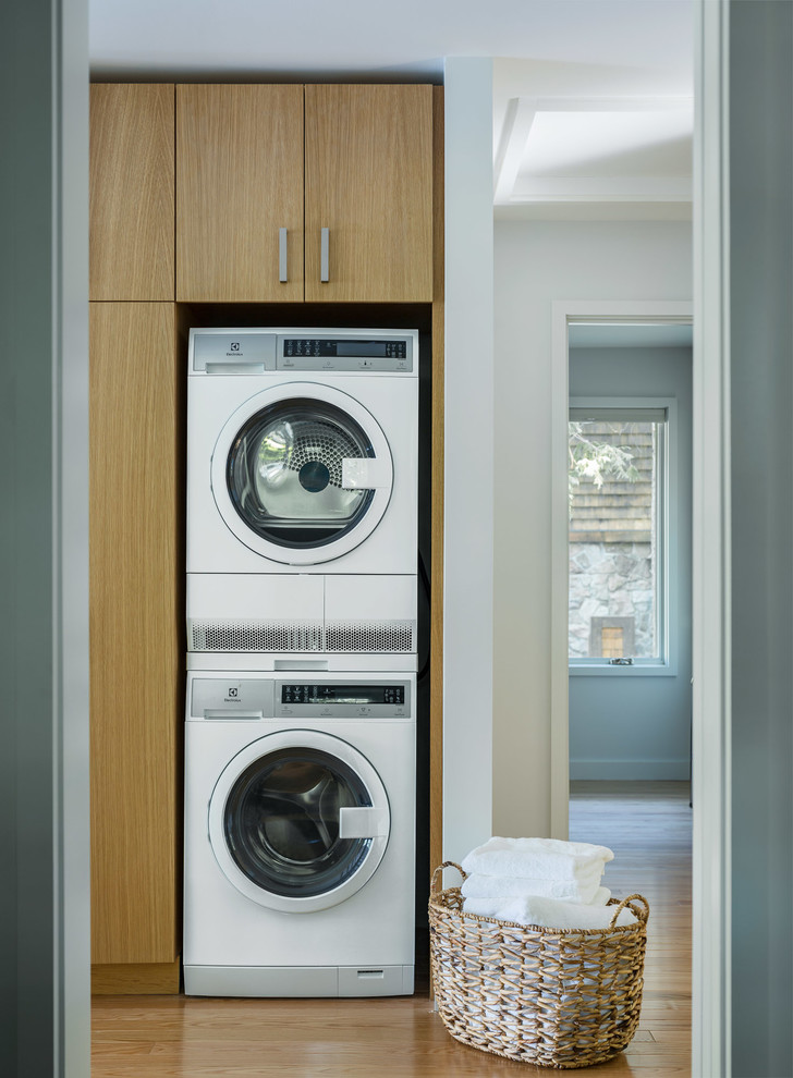 Design ideas for a small transitional single-wall dedicated laundry room in Burlington with flat-panel cabinets, light wood cabinets, medium hardwood floors, a stacked washer and dryer and brown floor.