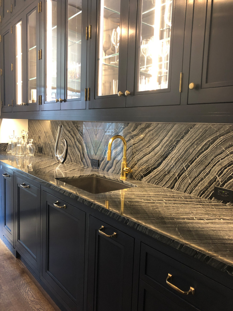Photo of an expansive transitional l-shaped kitchen pantry in New York with an undermount sink, recessed-panel cabinets, grey cabinets, marble benchtops, black splashback, stone slab splashback, panelled appliances, dark hardwood floors, brown floor and black benchtop.