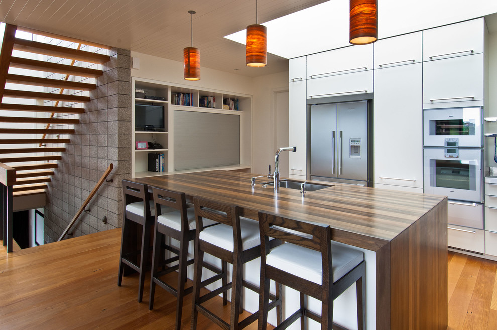 This is an example of a contemporary l-shaped kitchen in Wellington with an undermount sink, white cabinets, wood benchtops, stainless steel appliances, medium hardwood floors, with island and flat-panel cabinets.