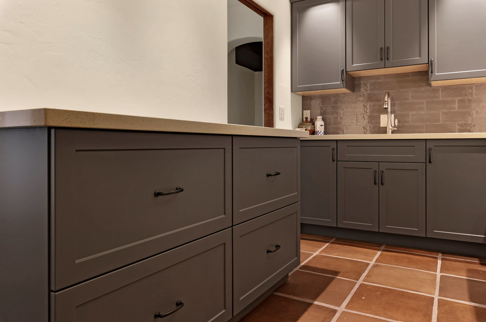 Design ideas for a mid-sized l-shaped dedicated laundry room in Phoenix with an undermount sink, shaker cabinets, grey cabinets, white walls, terra-cotta floors, a side-by-side washer and dryer, brown floor and white benchtop.