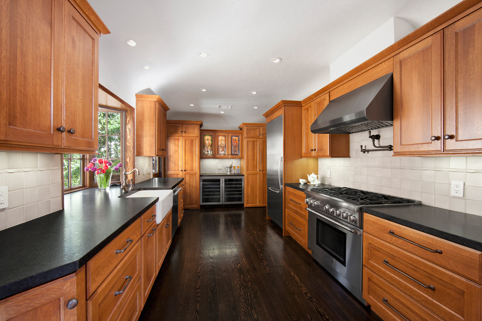 This is an example of a large transitional u-shaped separate kitchen in Los Angeles with a farmhouse sink, recessed-panel cabinets, medium wood cabinets, solid surface benchtops, grey splashback, stainless steel appliances, dark hardwood floors, no island, brown floor and black benchtop.